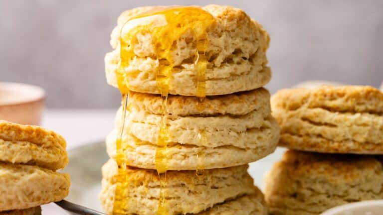 A stack of flaky biscuits with golden honey drizzled over the top, set on a plate with more biscuits in the background.