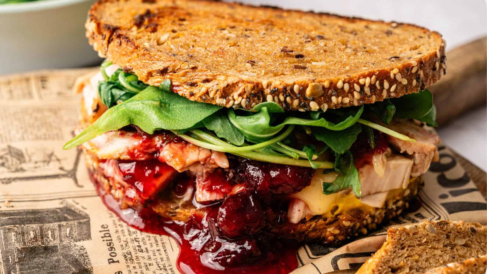 A sandwich made with whole grain bread, sliced turkey, melted cheese, leafy greens, and cranberry sauce sits on newspaper-patterned parchment.