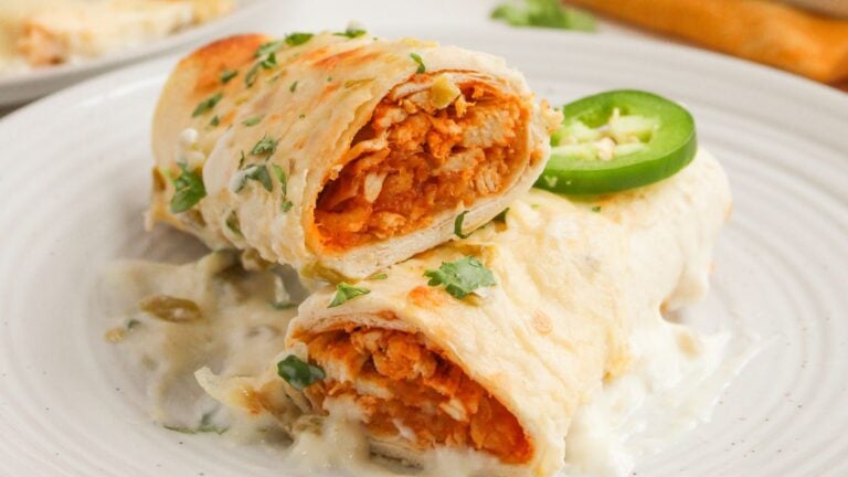 Two enchiladas topped with white sauce and garnished with sliced jalapeño and cilantro are served on a white plate. The filling appears to be shredded chicken.