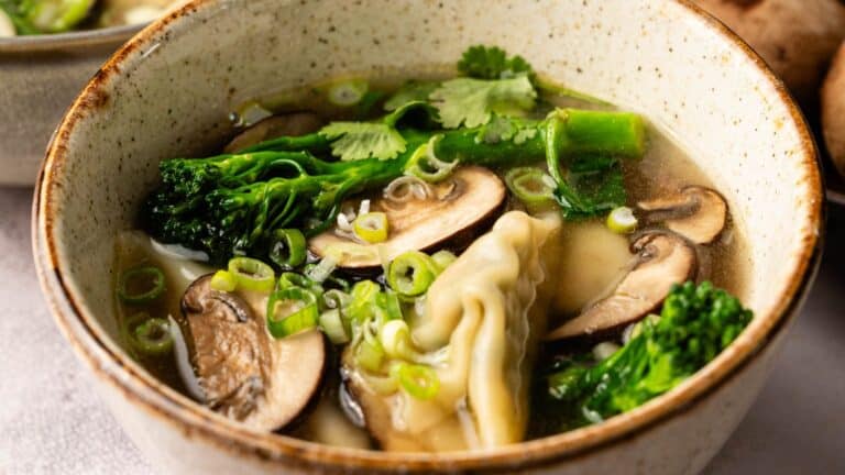A bowl of soup containing dumplings, sliced mushrooms, broccolini, green onions, and cilantro in a light broth.