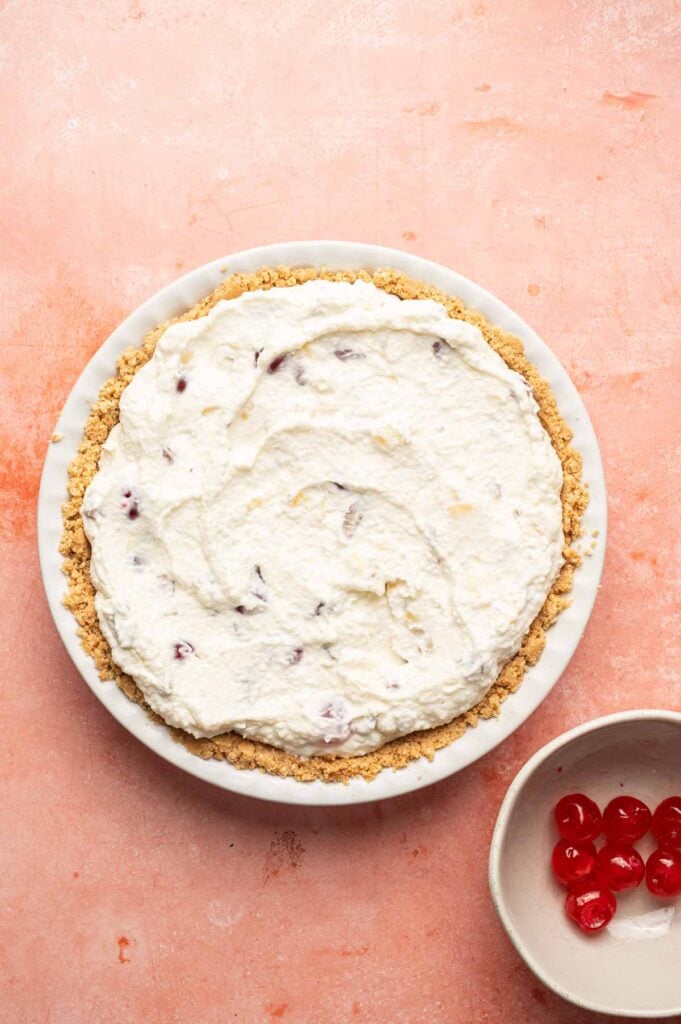 A Million Dollar Pie with a crumb crust and whipped cream topping sits on a pink surface next to a small bowl of maraschino cherries.