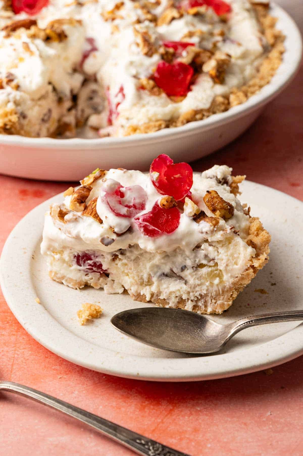 A slice of creamy Million Dollar Pie with chopped nuts and maraschino cherries on a white plate, a spoon beside it, and the remaining pie in the background.