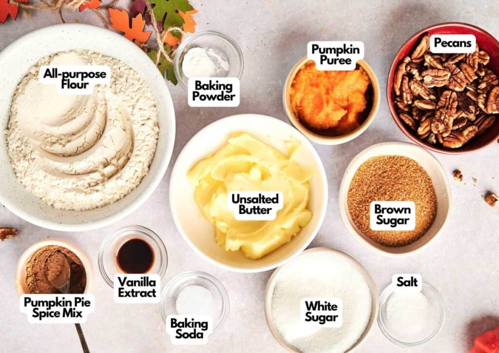 Top-down view of labeled baking ingredients for Air Fryer Pumpkin Cookies in bowls, including flour, baking powder, pumpkin puree, pecans, butter, brown sugar, white sugar, salt, baking soda, vanilla extract, and pumpkin pie spice mix.