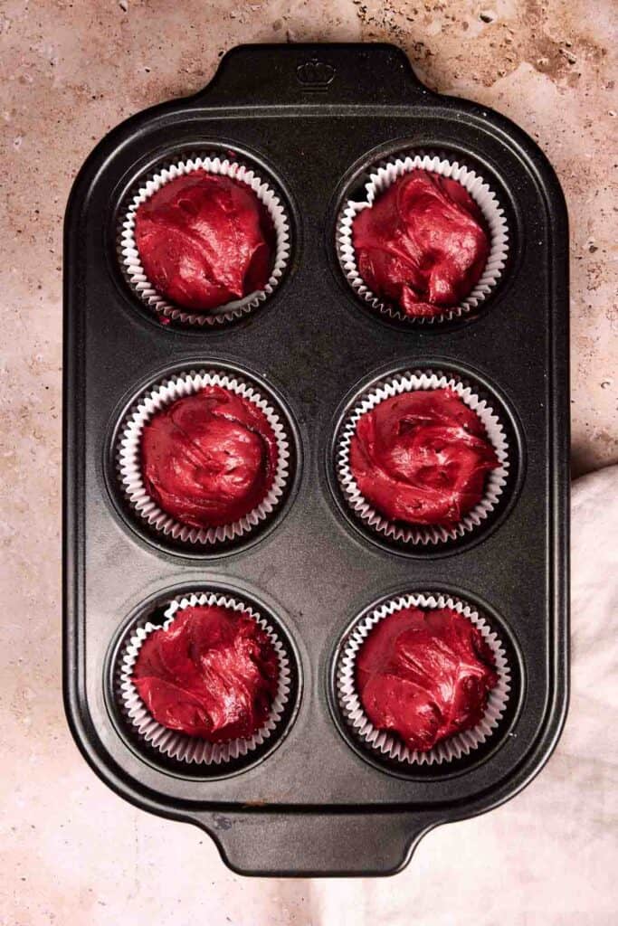 A black muffin tray with six paper liners is filled with Red Velvet Cupcakes batter, ready to bake on a speckled countertop.