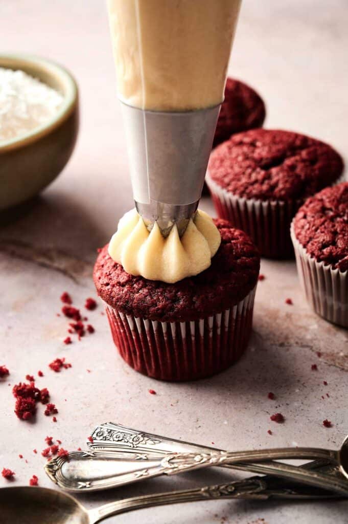 A piping bag frosts a red velvet cupcake, while unfrosted red velvet cupcakes, a bowl, and vintage spoons rest nearby on a marble surface.