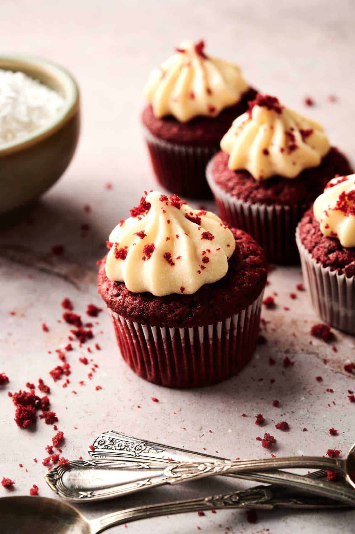 Three Red Velvet Cupcakes with cream cheese frosting and crumbs are arranged on a light surface near a bowl of shredded coconut and several silver forks.