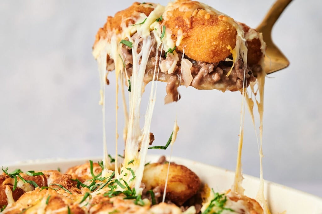 A serving utensil lifts a portion of cheesy Tater Tot Casserole with browned topping, ground meat, and fresh herbs, showing melted cheese stretching from the dish.