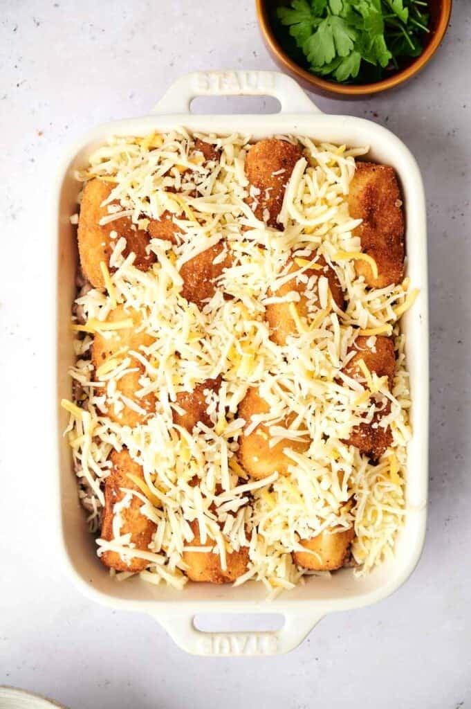 A rectangular white baking dish filled with breaded food pieces, resembling Tater Tot Casserole, topped with shredded cheese, next to a bowl of fresh parsley.