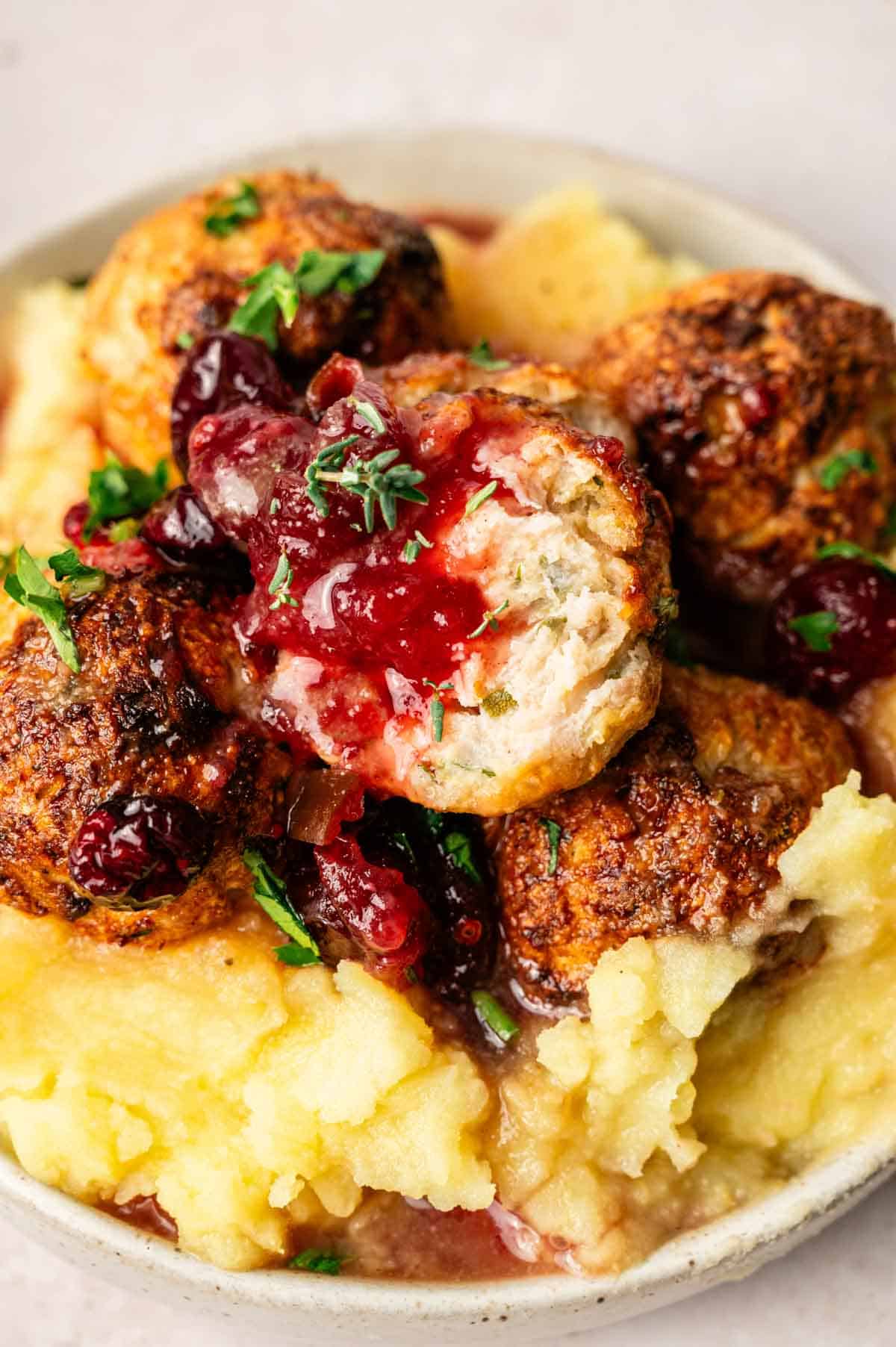 A plate of mashed potatoes topped with browned Thanksgiving Meatballs, cranberry sauce, and chopped herbs.