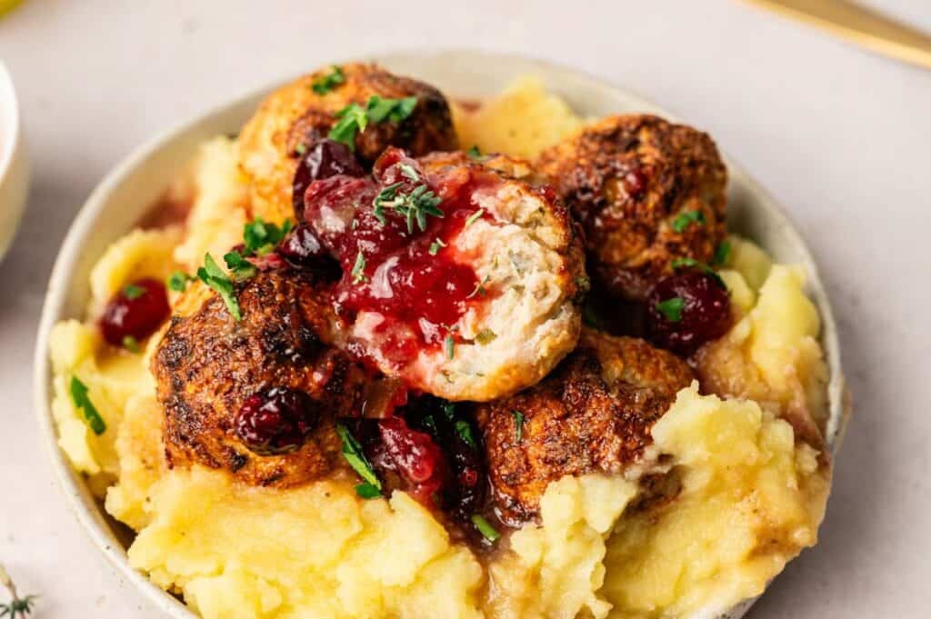 A bowl of mashed potatoes topped with browned Thanksgiving Meatballs, red cranberry sauce, and garnished with chopped parsley.