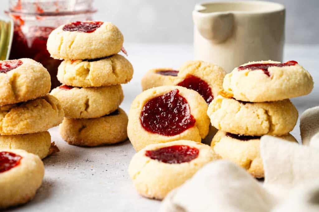Stacks of Thumbprint Cookies filled with red jam are arranged on a light surface, with a jar of jam and a ceramic container in the background.