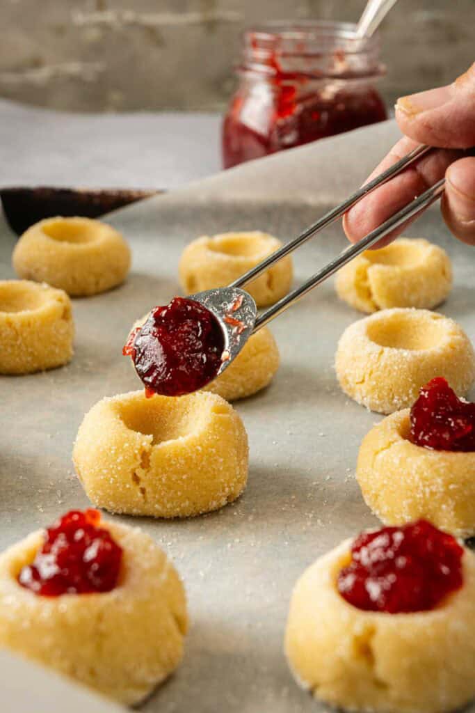 A hand uses a spoon to fill indented rounds of raw Thumbprint Cookie dough with red jam on a parchment-lined baking sheet; a jar of jam is in the background.