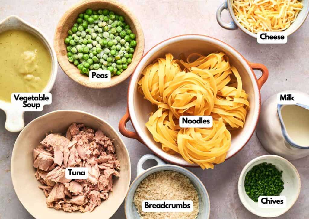 Various ingredients for a Tuna Casserole are arranged in bowls, including vegetable soup, peas, noodles, cheese, milk, tuna, breadcrumbs, and chives.