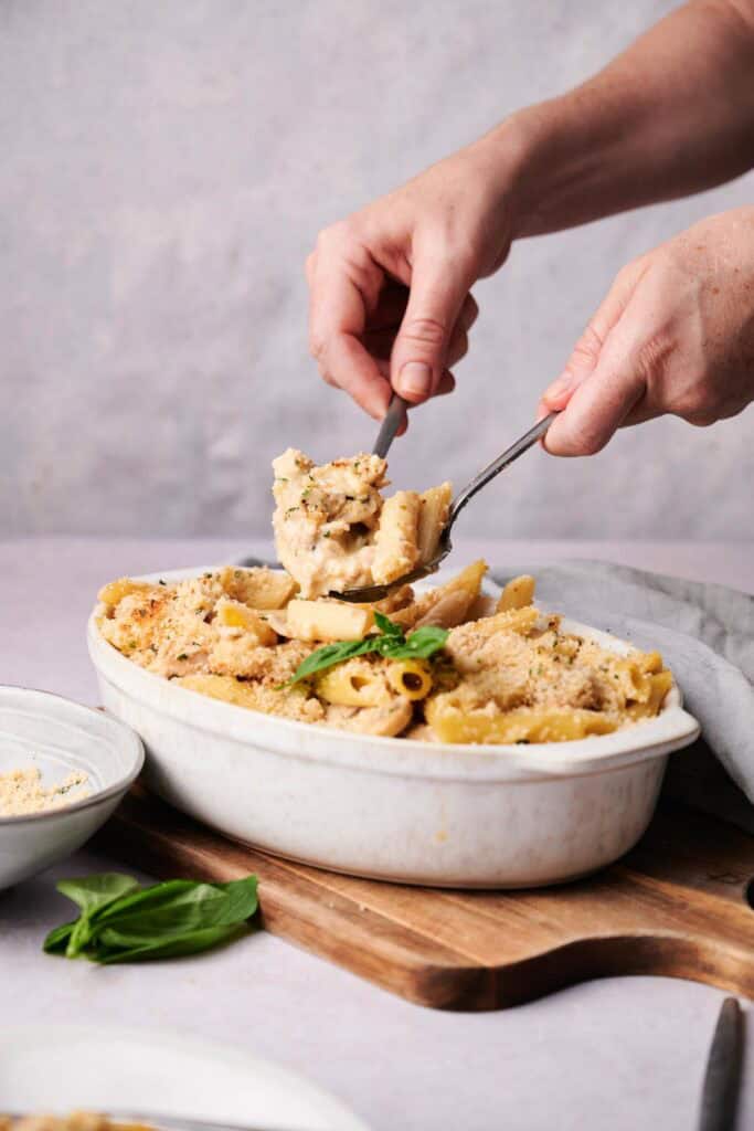 Person serving a portion of Turkey Casserole baked pasta from a white ceramic dish, garnished with basil, onto a plate.