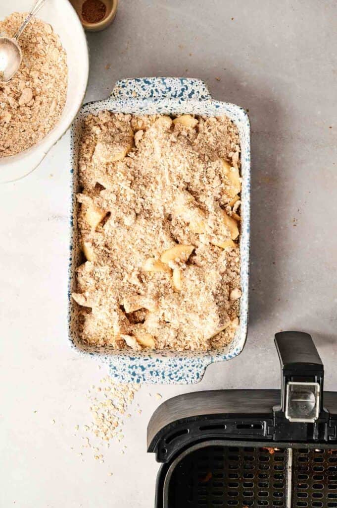 A baking dish of Air Fryer Apple Crisp with sliced apples and crumb topping sits by an air fryer basket and a bowl of extra topping on a light countertop.