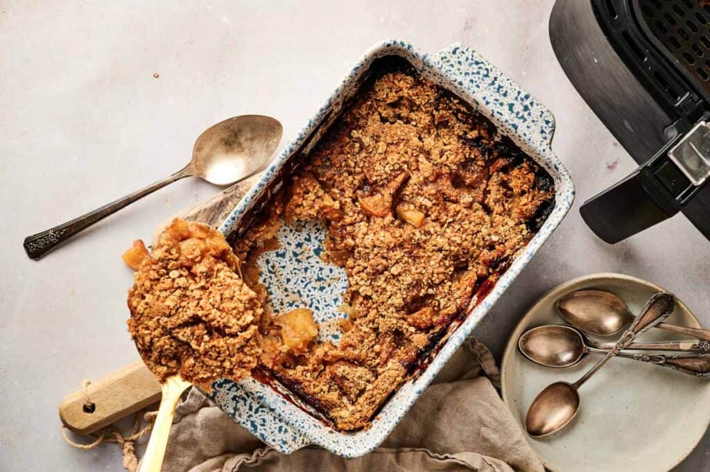 A rectangular baking dish with Air Fryer Apple Crisp, a large serving spoon, and several small spoons and plates on the side.