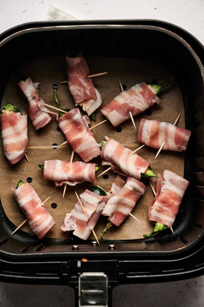 Air Fryer Bacon Wrapped Jalapeno Poppers, secured with toothpicks, are neatly arranged on parchment paper in the air fryer basket, ready to cook.