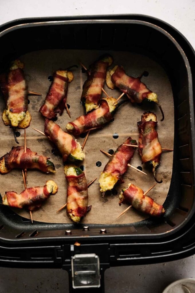 Air Fryer Bacon Wrapped Jalapeno Poppers are arranged on parchment paper in the basket, secured with toothpicks and perfectly cooked for a crispy, savory bite.