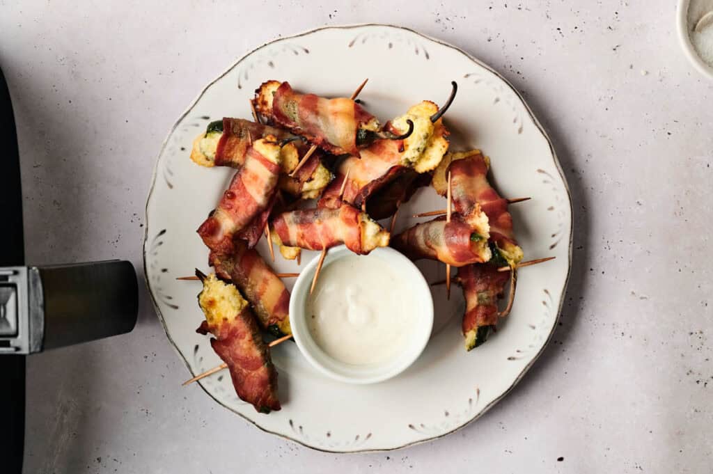 A plate of Air Fryer Bacon Wrapped Jalapeno Poppers encircles a small bowl of white dipping sauce in the center.