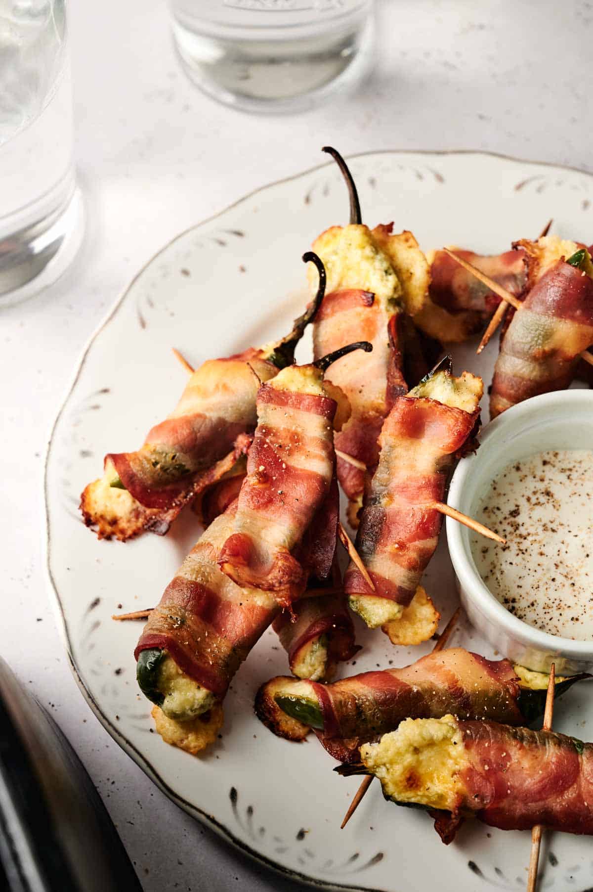 A plate of Air Fryer Bacon Wrapped Jalapeno Poppers secured with toothpicks sits next to a small bowl of creamy dipping sauce and two glasses of water.