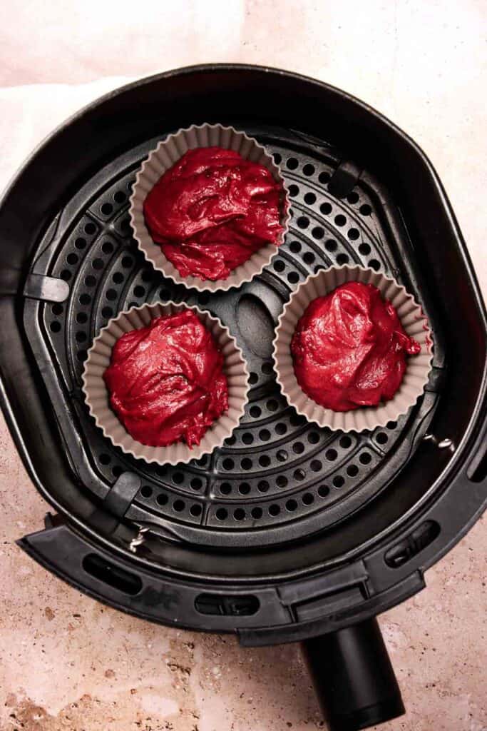 Three silicone cupcake liners filled with red batter are arranged inside an air fryer basket, ready to become delicious Air Fryer Red Velvet Cupcakes.