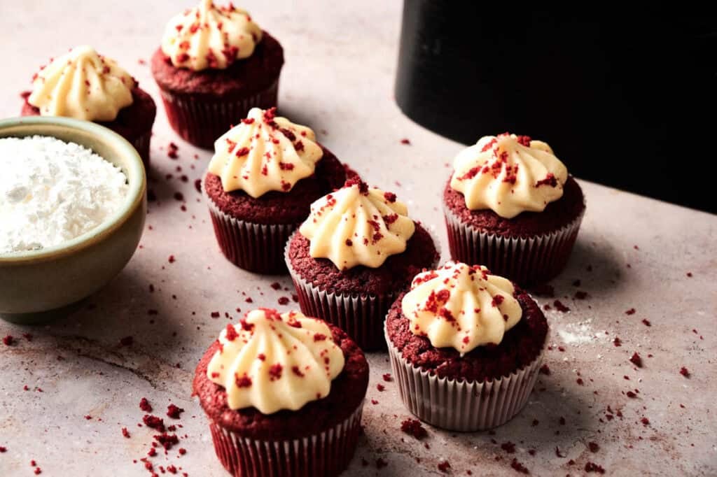 Six Air Fryer Red Velvet Cupcakes with cream cheese frosting, topped with red crumbs, are arranged on a light surface next to a bowl of powdered sugar.