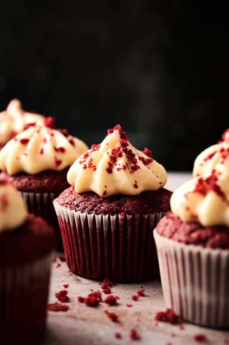 Air Fryer Red Velvet Cupcakes with cream cheese frosting and red cake crumbs on top, arranged on a light surface with a dark background.