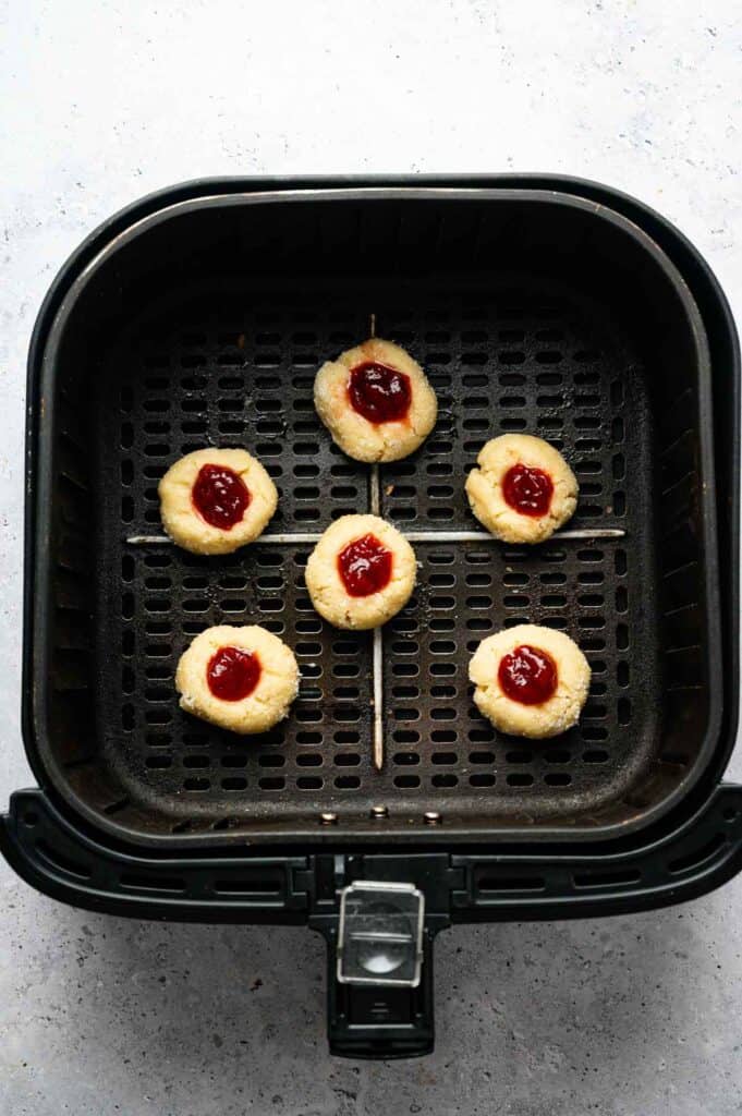 Six unbaked Air Fryer Thumbprint Cookies with red jam centers are arranged neatly in an air fryer basket, ready to bake.