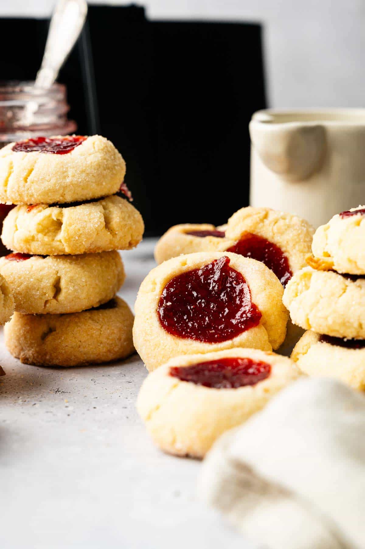 Stacks of Air Fryer Thumbprint Cookies filled with red jam are arranged on a light surface, with a jar and a ceramic pitcher in the background.