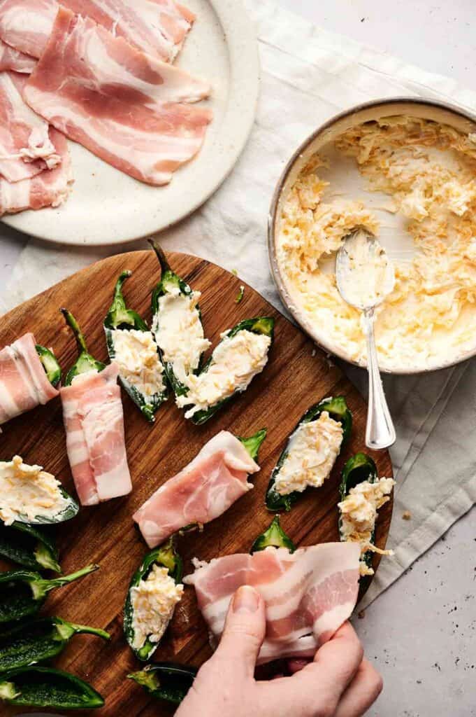 A hand wraps bacon around jalape&ntilde;o halves stuffed with cheese mixture on a wooden board; Air Fryer Bacon Wrapped Jalapeno Poppers and ingredients are nearby.