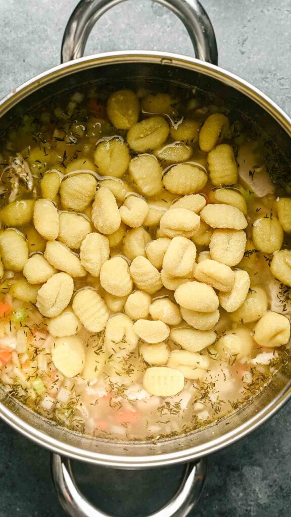 A stainless steel pot filled with flavorful Gnocchi Soup, topped with uncooked gnocchi pieces floating on the surface.