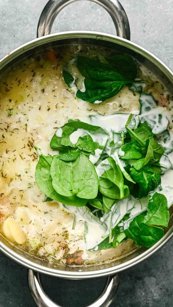 A stainless steel pot containing creamy Gnocchi Soup with fresh spinach leaves and herbs on top, ready to be stirred.