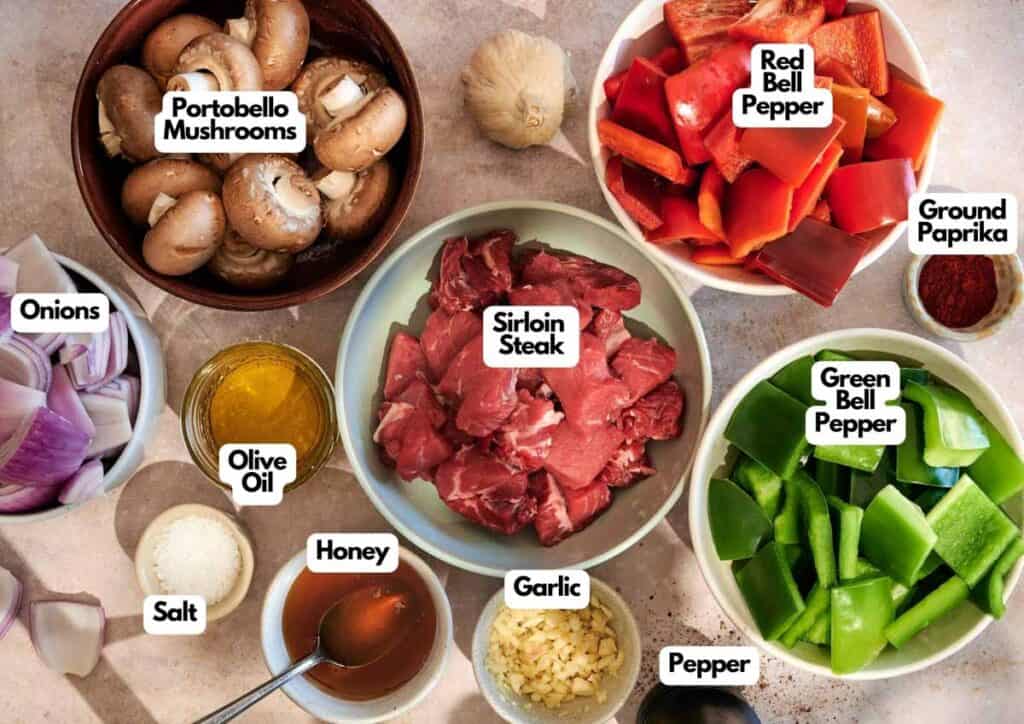 A top-down view of bowls containing sirloin steak for Beef Kabobs with Chimichurri Sauce, portobello mushrooms, red and green bell peppers, onions, garlic, olive oil, honey, salt, pepper, and ground paprika.