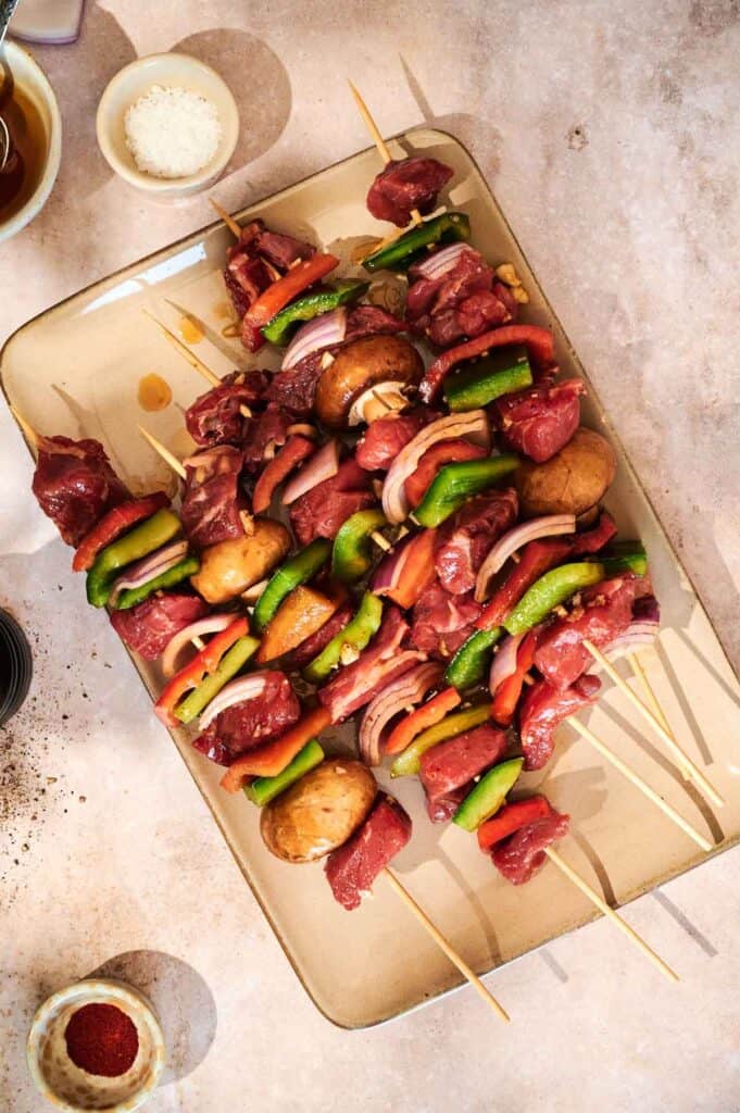 Raw beef kabobs with green peppers, red peppers, onions, and mushrooms on wooden skewers are arranged on a beige tray—perfect for pairing with zesty chimichurri sauce.