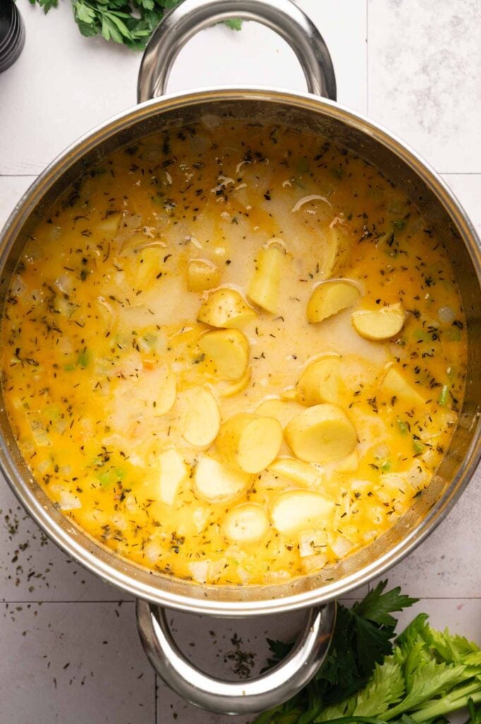 A pot of sliced potatoes and herbs simmering in a yellow broth, viewed from above on a light countertop, with some fresh herbs nearby—an inviting scene perfect for crafting a comforting Ham and Potato Soup.