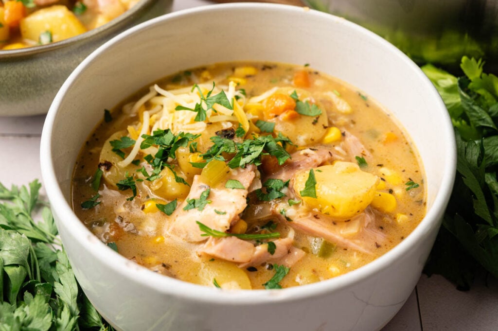 A bowl of creamy chicken and vegetable soup, reminiscent of comforting Ham and Potato Soup, is garnished with chopped parsley and shredded cheese, surrounded by fresh parsley.