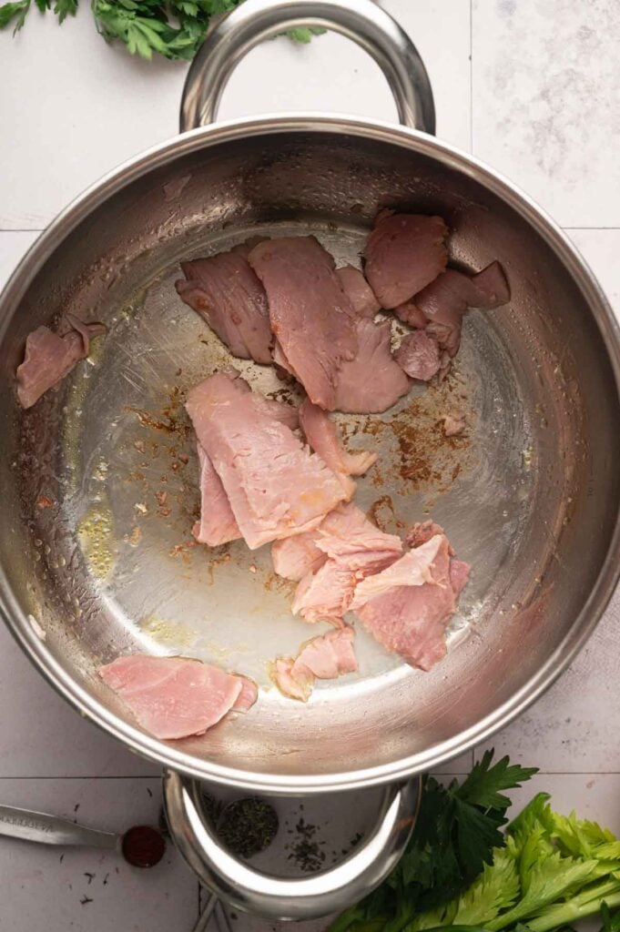Stainless steel pot with slices of ham being cooked, some browning visible on the surface; fresh herbs are partially visible around the pot, suggesting the savory beginnings of a homemade Ham and Potato Soup.