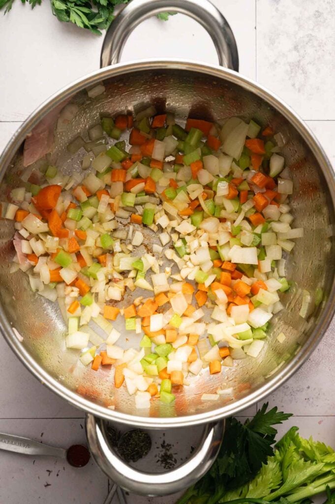 Diced onions, carrots, celery, and garlic are being sautéed in a stainless steel pot as the delicious base for Ham and Potato Soup, with herb sprigs and leafy greens visible around the pot on a countertop.