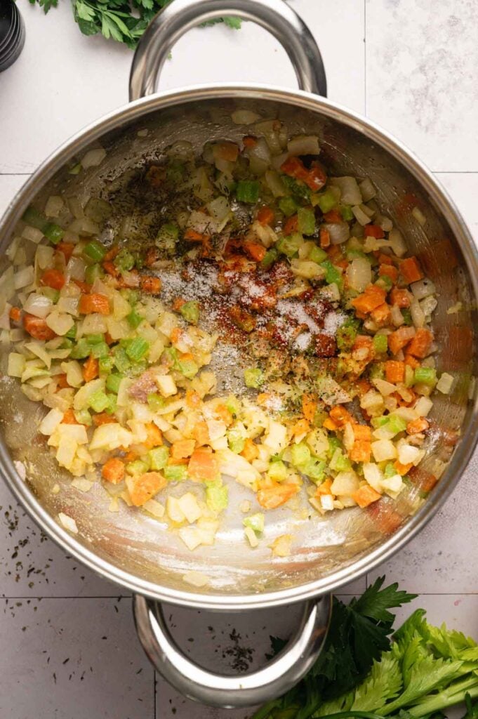 Diced onions, carrots, and celery sauté in a large pot with spices sprinkled on top—the flavorful base for Ham and Potato Soup; fresh herbs and celery are nearby on the counter.