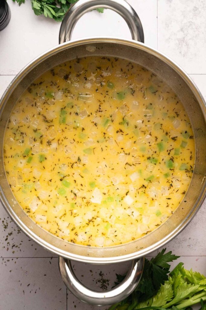 A pot of Ham and Potato Soup with chopped onions, celery, hearty potatoes, savory broth, and fresh herbs simmering on a stovetop, viewed from above.