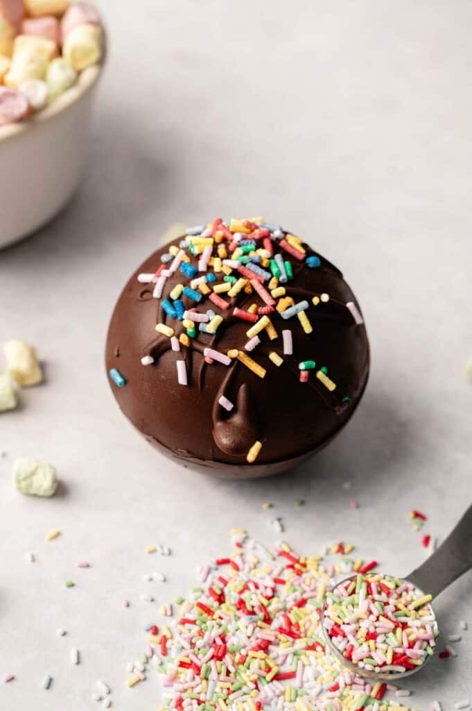 A round Hot Chocolate Bomb topped with colorful sprinkles sits on a light surface, surrounded by extra sprinkles and mini marshmallows, ready to create a deliciously sweet treat.