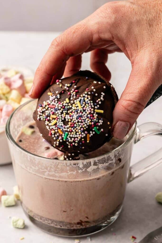 A hand dips a chocolate-covered Hot Chocolate Bomb with colorful sprinkles into a glass mug of hot chocolate; marshmallows are visible in the background.