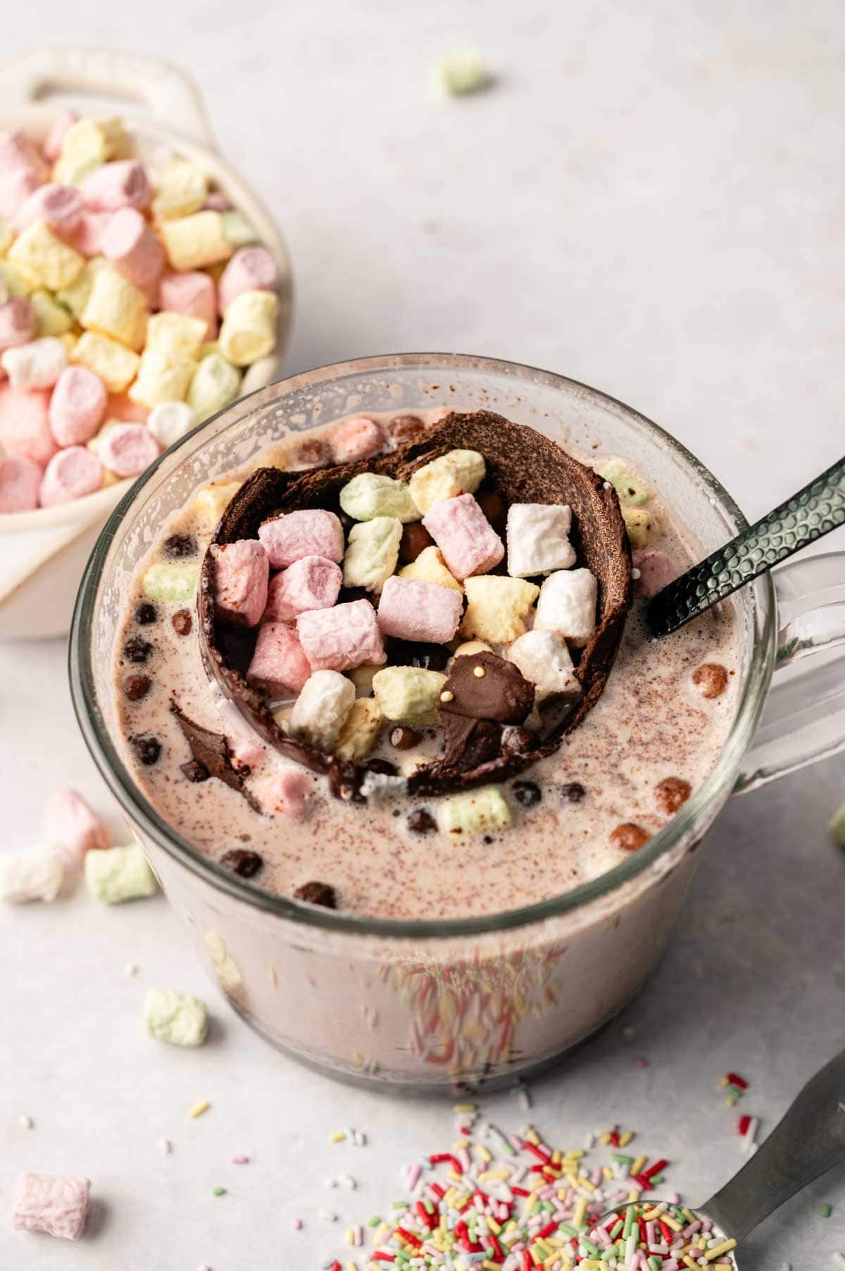 A glass mug of hot chocolate with a Hot Chocolate Bomb and colorful mini marshmallows on top, surrounded by extra marshmallows and sprinkles in the background.