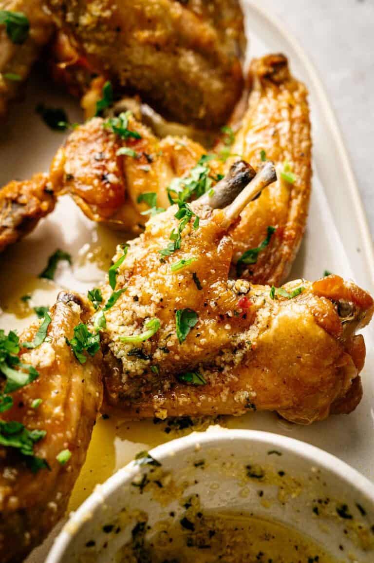 Close-up of baked Lemon Pepper Wings garnished with chopped herbs and grated cheese, with a bowl of sauce in the foreground.
