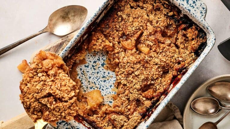 A baked apple crumble in a blue speckled dish, with a serving spoon removing a portion; some apple filling and crumb topping are visible.