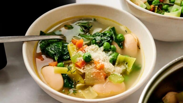 A bowl of vegetable soup with spinach, peas, carrots, celery, and large white beans, topped with grated cheese, with a spoon inside the bowl.