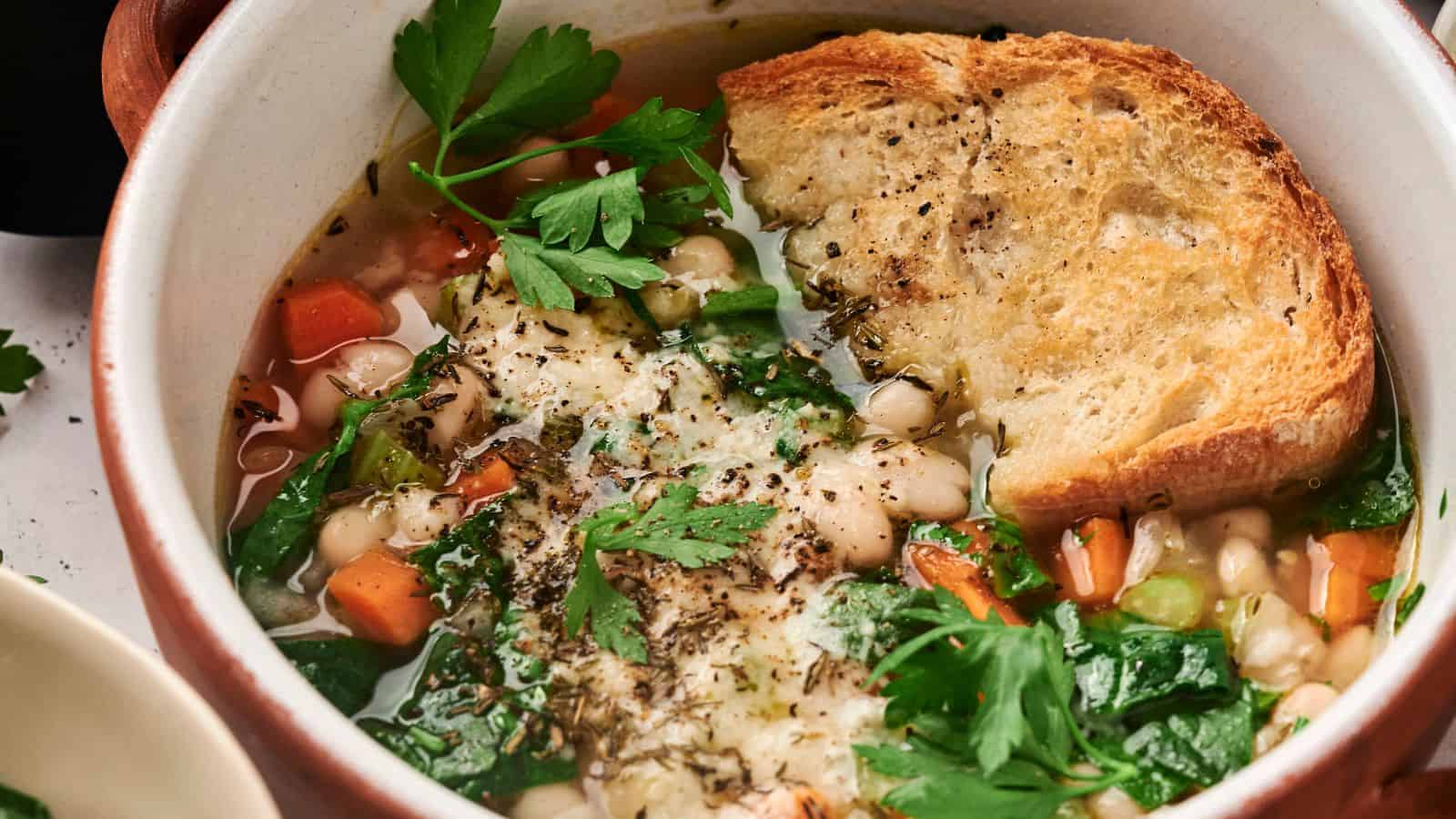 A bowl of vegetable and white bean soup is topped with parsley, grated cheese, and a slice of toasted bread.