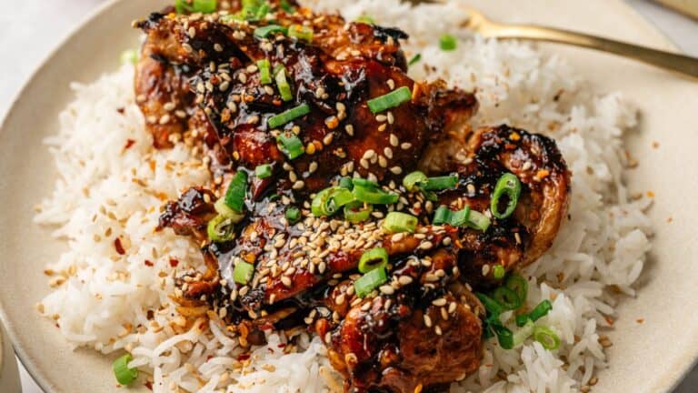 A plate of white rice topped with glazed chicken, garnished with sliced green onions and sesame seeds.