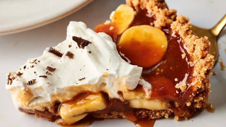 A slice of banoffee pie with a crumbly crust, caramel, banana slices, whipped cream, and chocolate shavings on top, served on a white plate.
