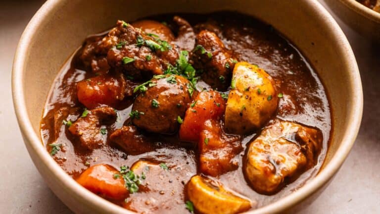 A bowl of beef stew with chunks of beef, potatoes, carrots, and mushrooms in a thick brown gravy, garnished with chopped parsley.